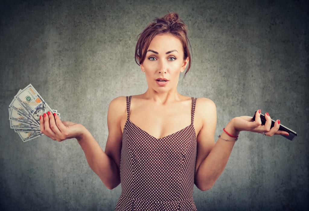 Woman shrugging shoulders with money in one hand and smartphone in other.