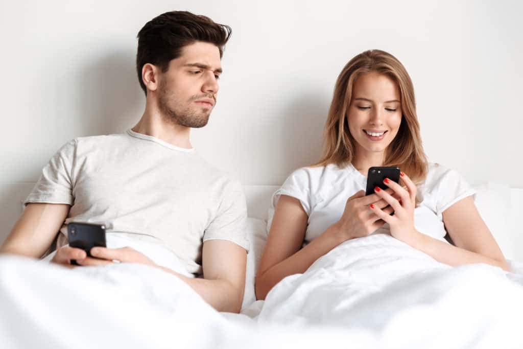 Couple in bed with man looking worriedly at woman's phone wondering are sms text messages secure and private