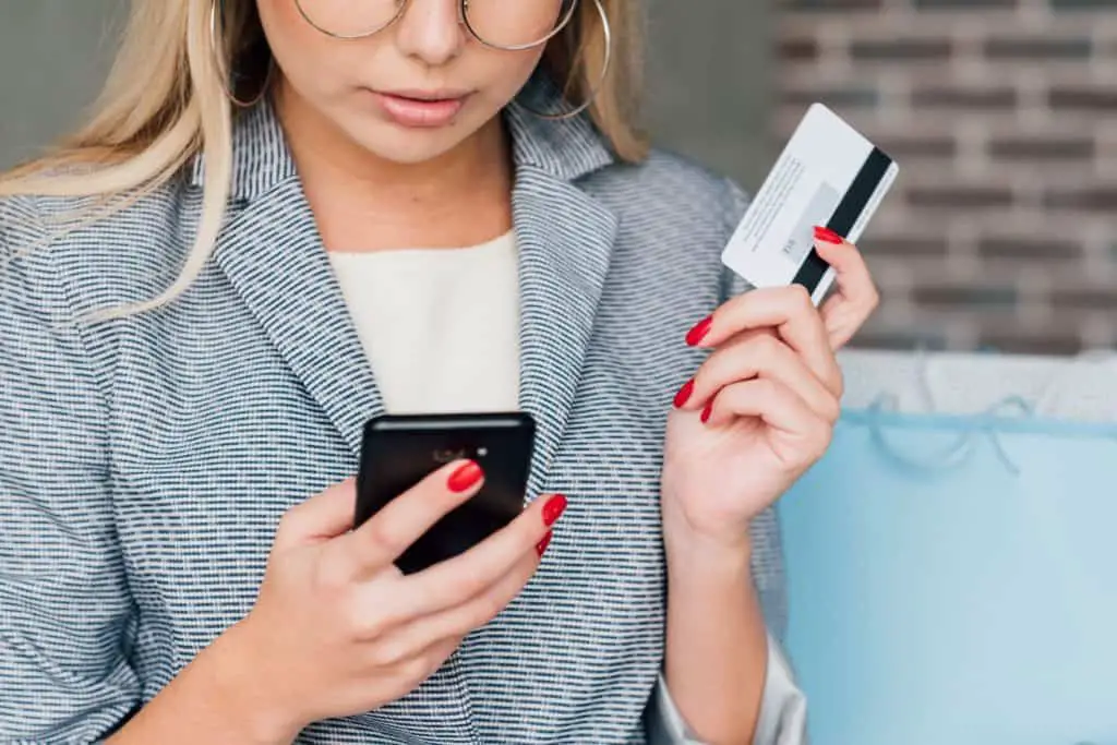 Woman with debit card and smartphone wondering if is it safer to use an app or browser for banking