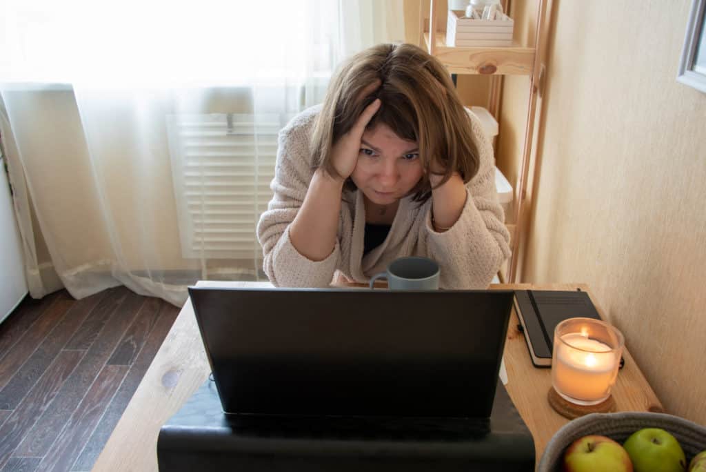 Woman with laptop at home with hand on head staring at laptop having just opened a spam email