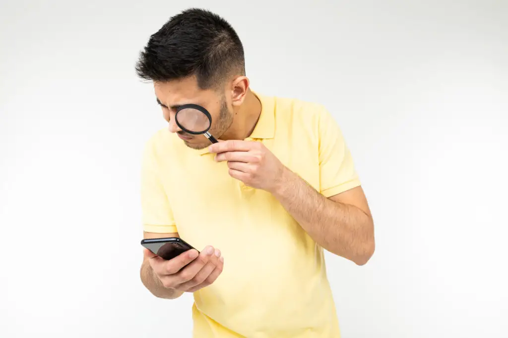 Man looking at his smartphone through a magnifying glass trying to see fake profile pictures