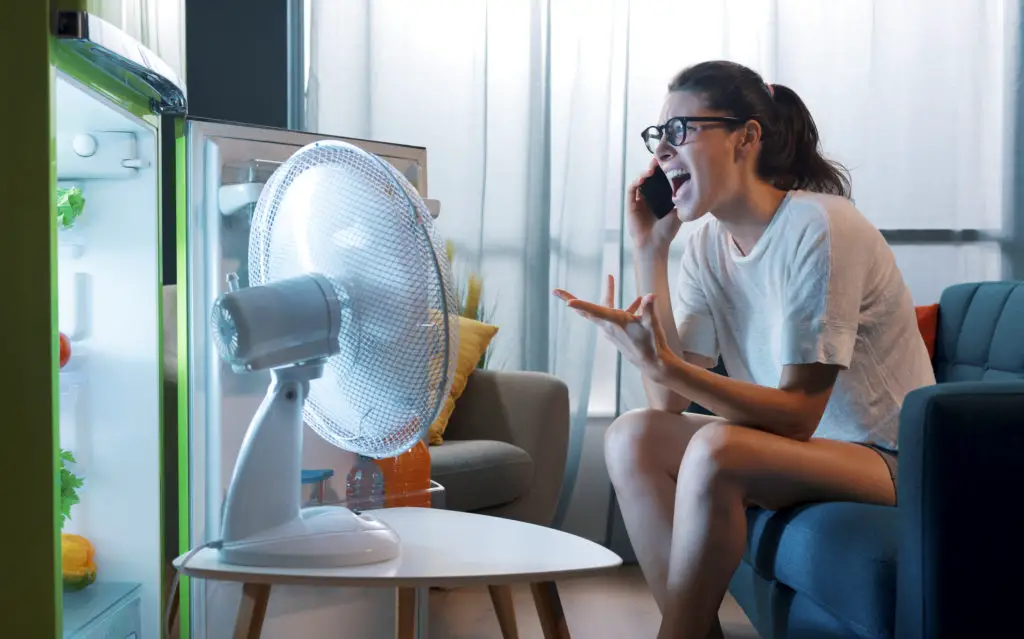 Woman on couch in front of a fan and open refigerator asking "why is my phone hot? "