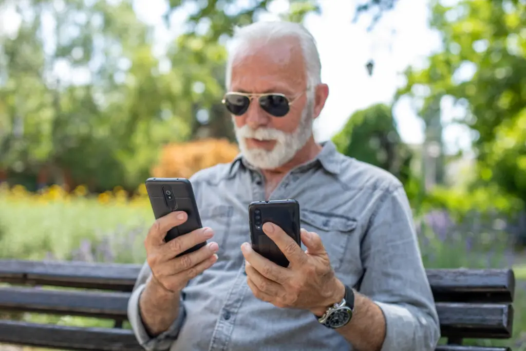 Man on a park bench with two smartphones wondering if a factory reset will protect against hackers