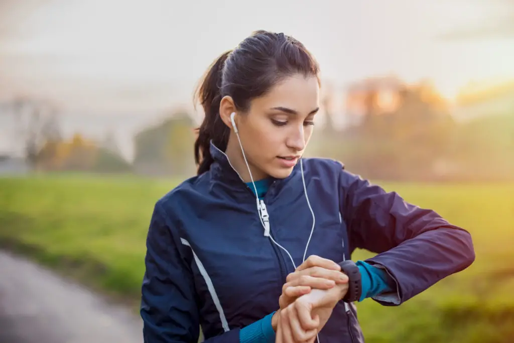 Woman with earbuds in pausing on her run to check her smartwatch.