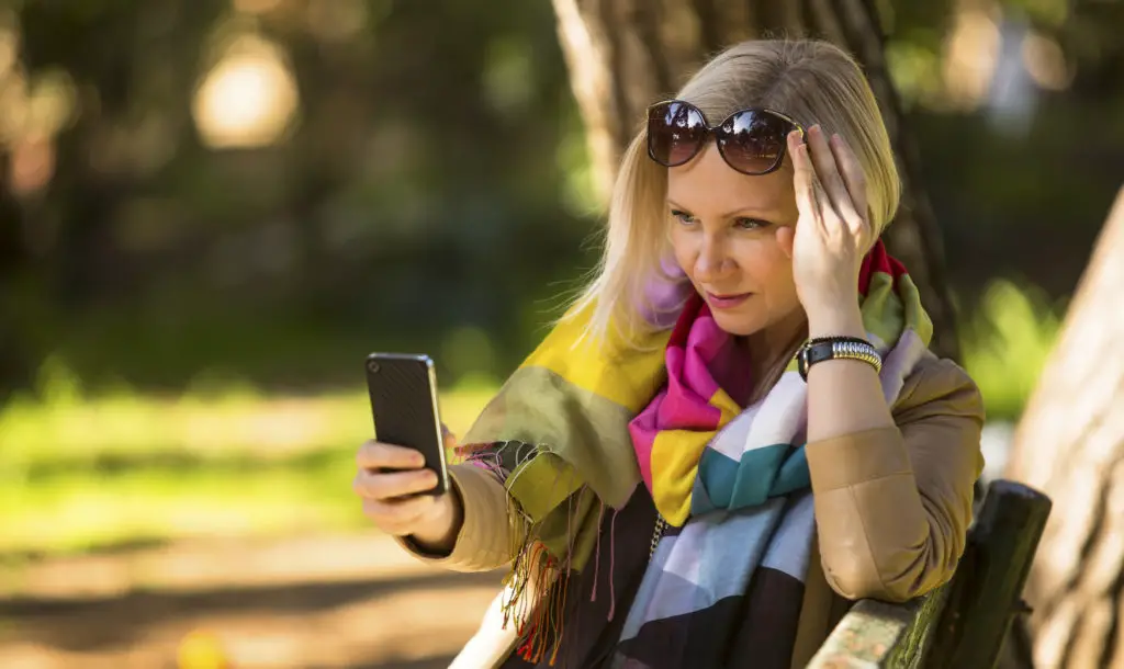 Woman with sunglasses in a park looking at her android phone with a confused expression because someone "liked" a text message.