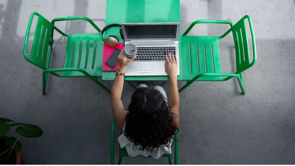 Unidentifiable woman sitting on a green table using laptop, holding coffee on one hand