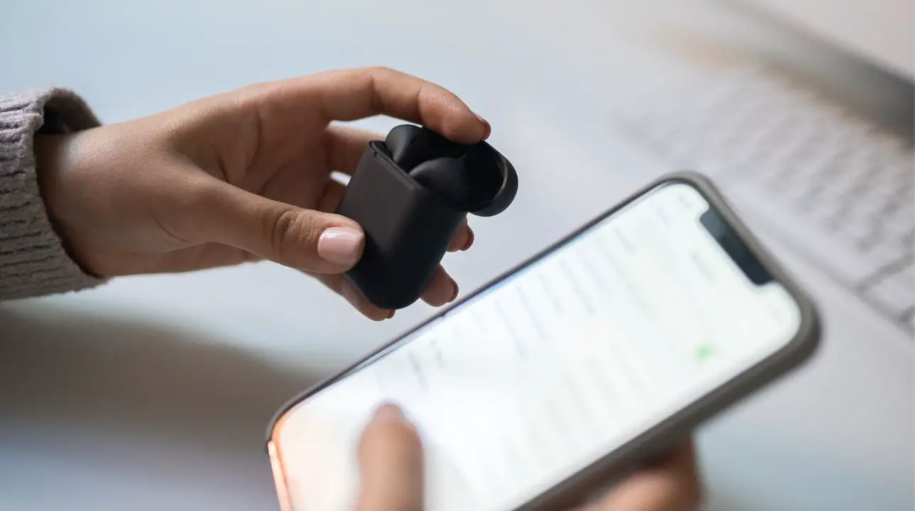 A person holding bluetooth wireless earbuds on one hand and a smartphone on the other