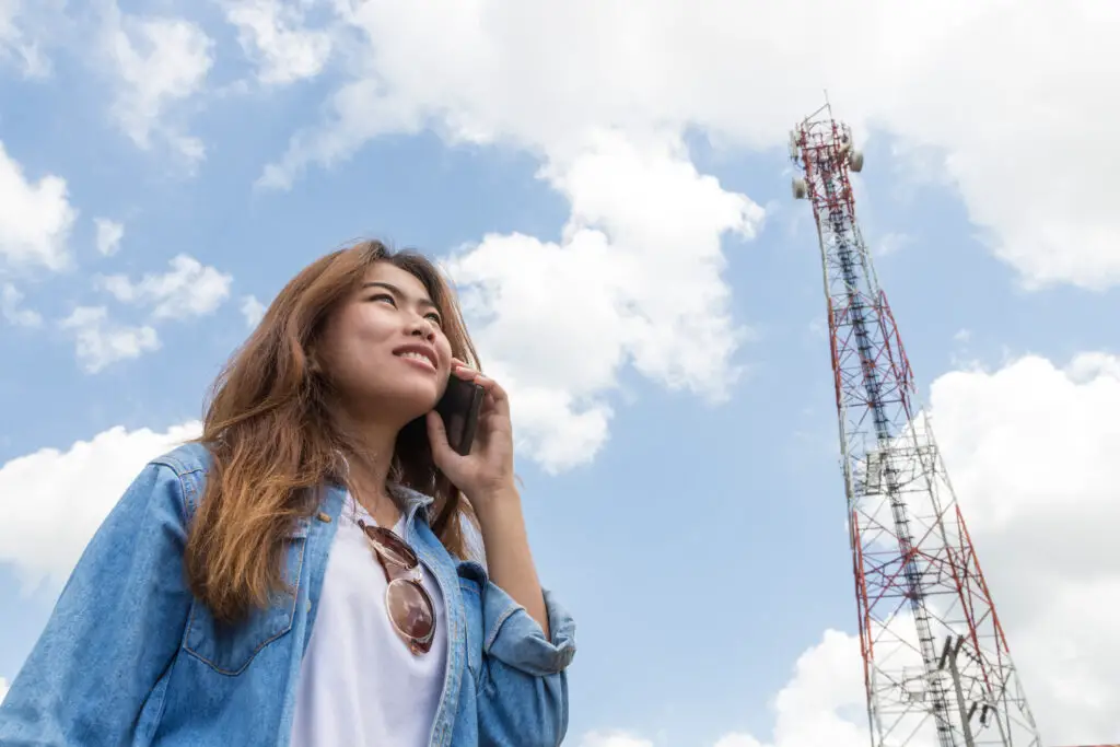 Woman on her smartphone next to a cellular tower wondering how mobile phones communicate
