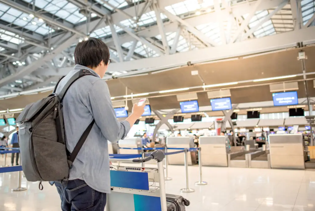 Man at airport ticket counter holding smartphone wondering can you put mobile phones in checked baggage