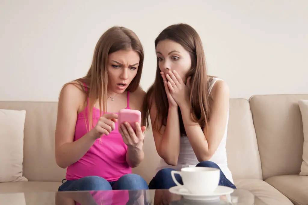 Two shocked young girls looking at a smartphone screen after having received inappropriate text messages.